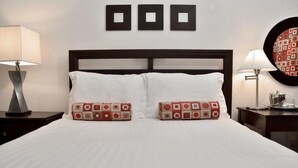 Un lit bien fait, avec du linge de lit blanc et deux coussins décoratifs, une tête de lit en bois foncé et un mur orné de trois cadres carrés.
