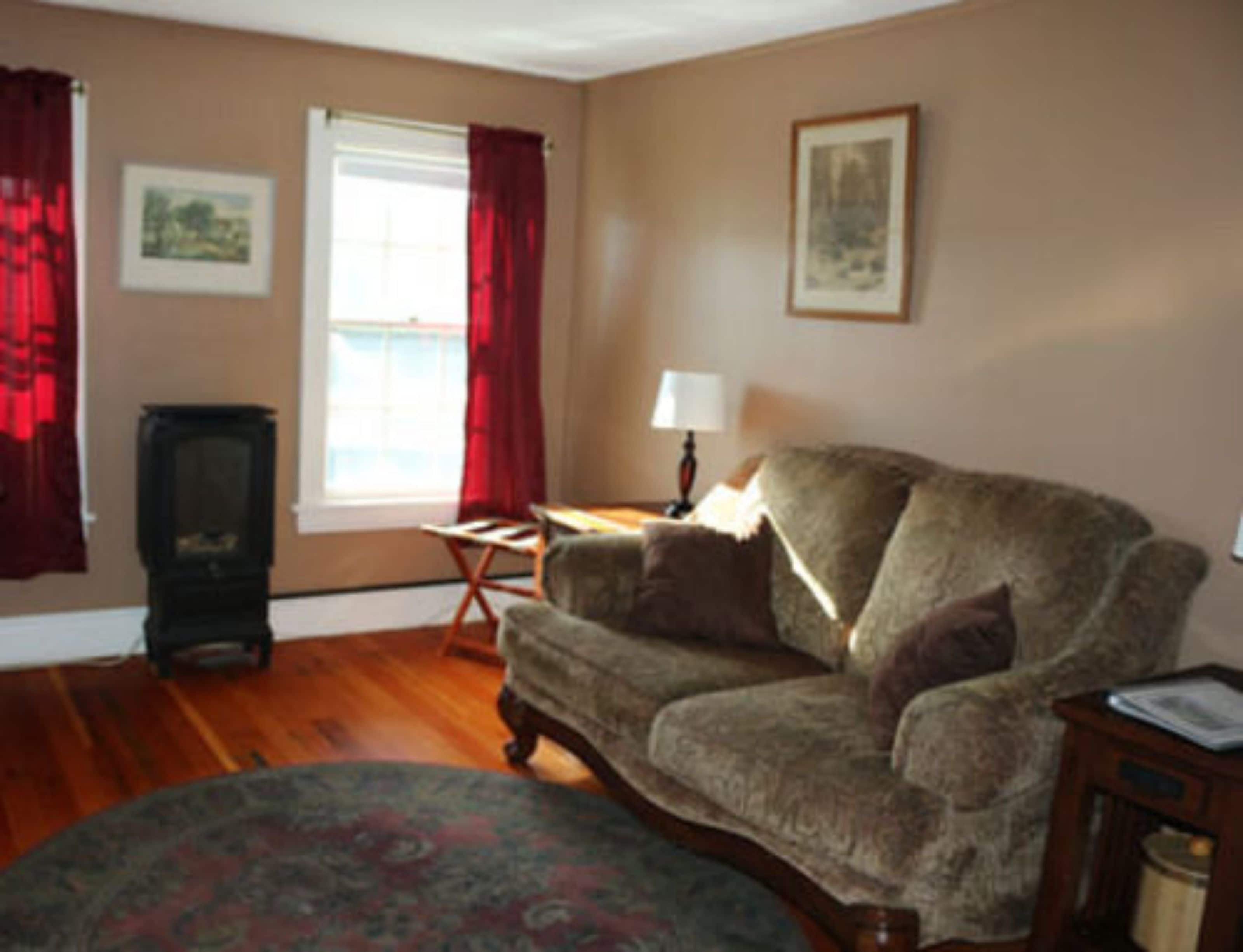 A living room with a wooden floor, a patterned rug, a sofa, a small table, a lamp, and a window with red curtains.
