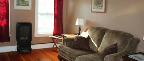 A living room with a wooden floor, a patterned rug, a sofa, a small table, a lamp, and a window with red curtains.