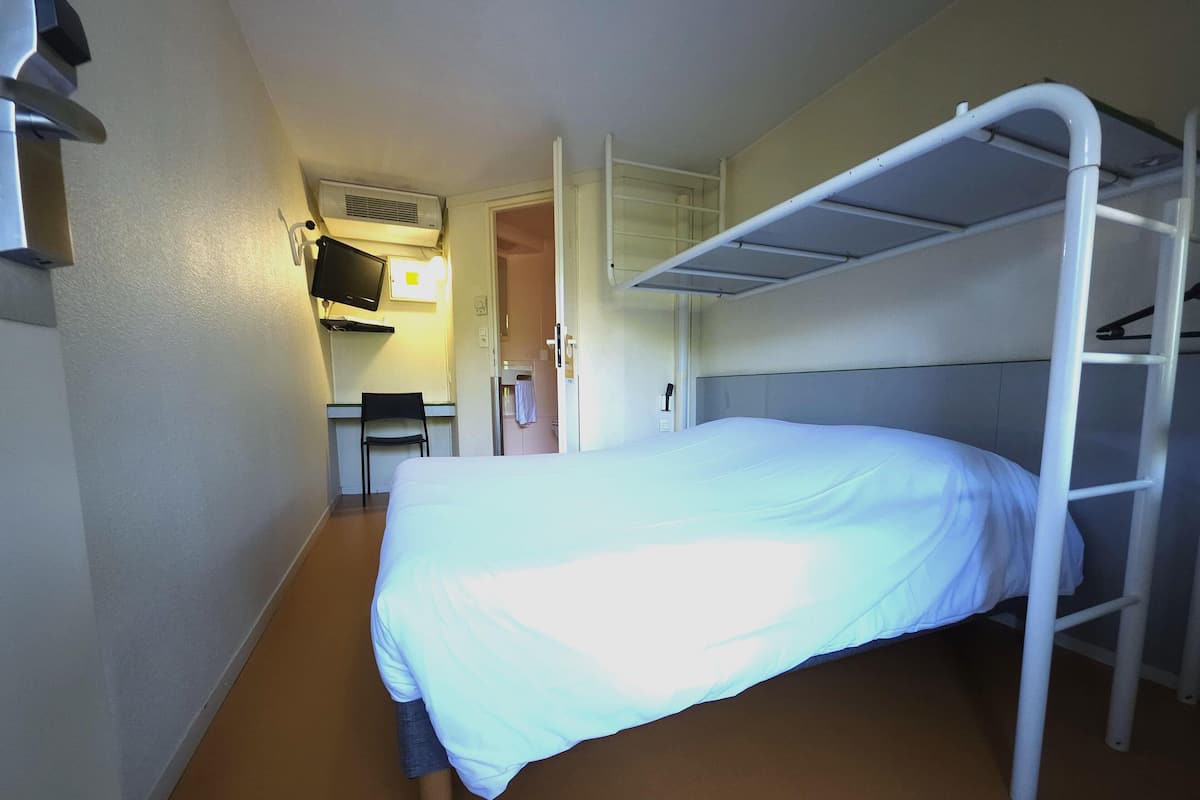 Quarto de hotel compacto com beliche, escrivaninha com cadeira, banheiro pequeno e televisão montada na parede.