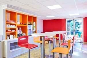 Une cafétéria lumineuse, avec des meubles aux couleurs orange et blanche, un comptoir où sont exposés divers articles, et une vue sur la verdure à l’extérieur.