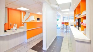A brightly lit reception area with orange and white decor, a large white logo on the wall, and a counter with a phone and informational materials.