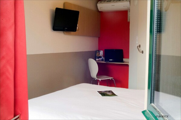 Um quarto com uma cama, uma cadeira branca, uma escrivaninha com um laptop, uma televisão na parede e uma janela com vista.