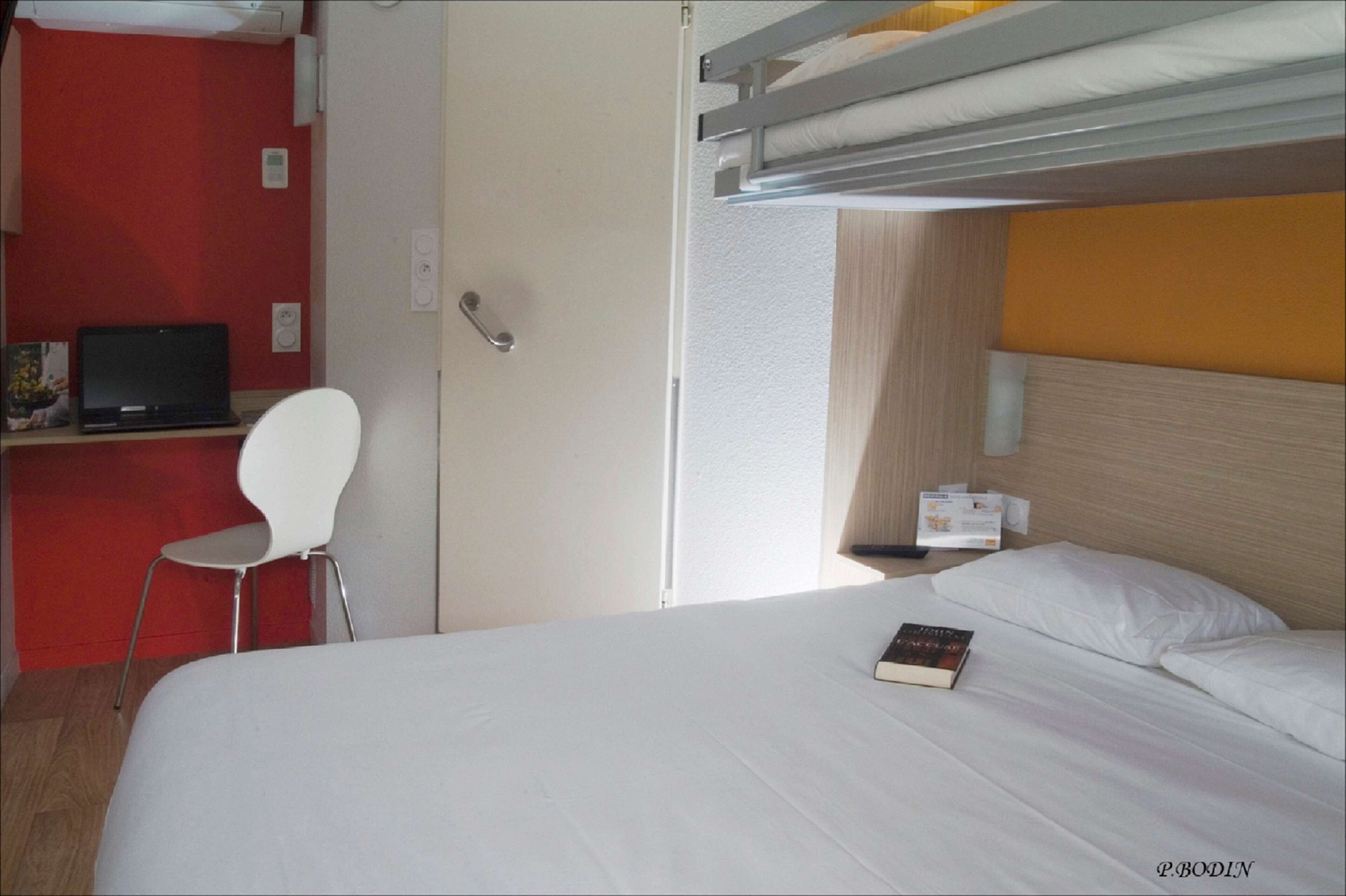 A hotel room with a bunk bed, a desk with a laptop, a white chair, and a bedside table with a book.