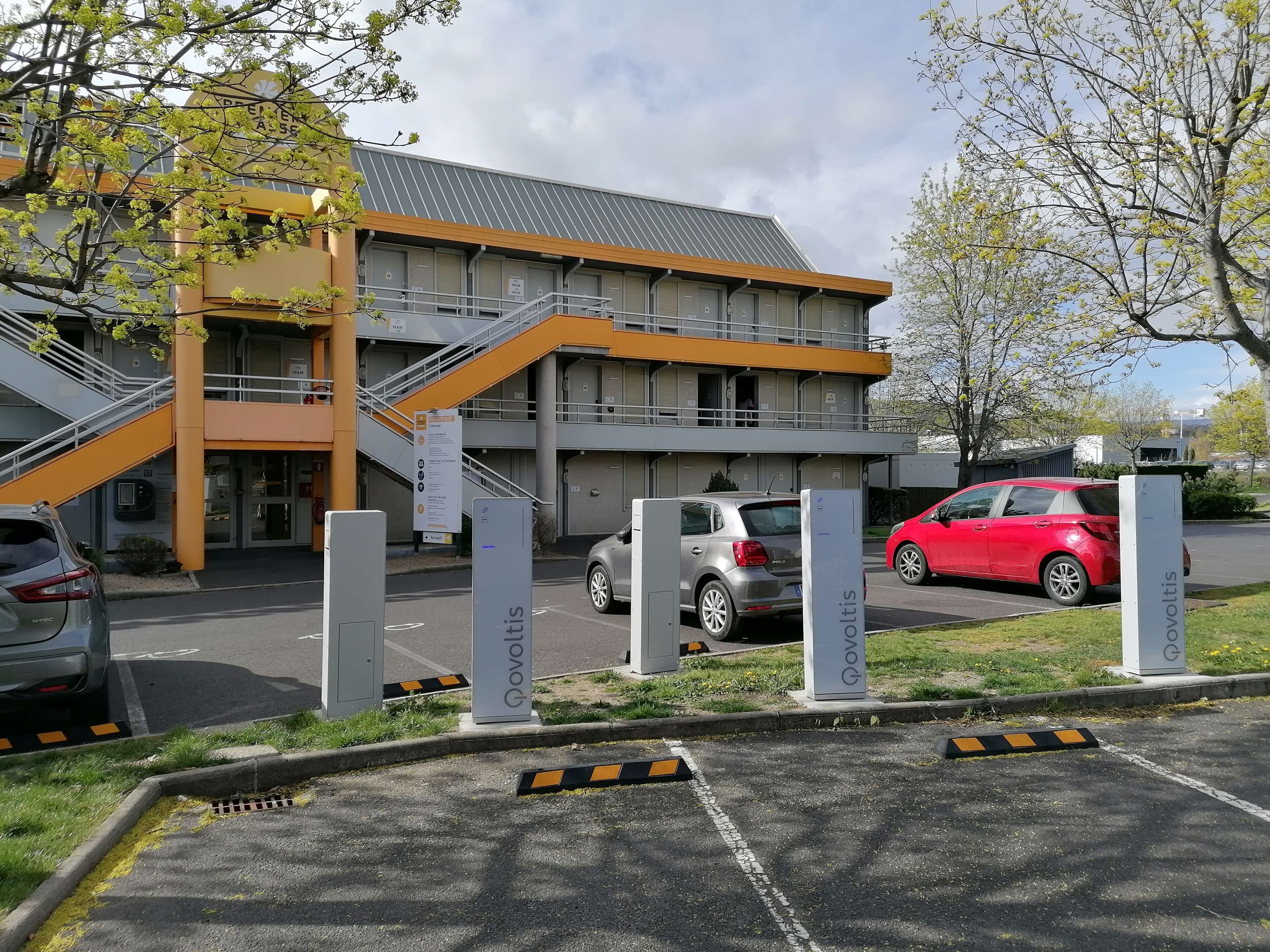 A building with a modern design, featuring a staircase leading to an upper level. There are parking spaces in front of the building, and a red car is parked nearby.