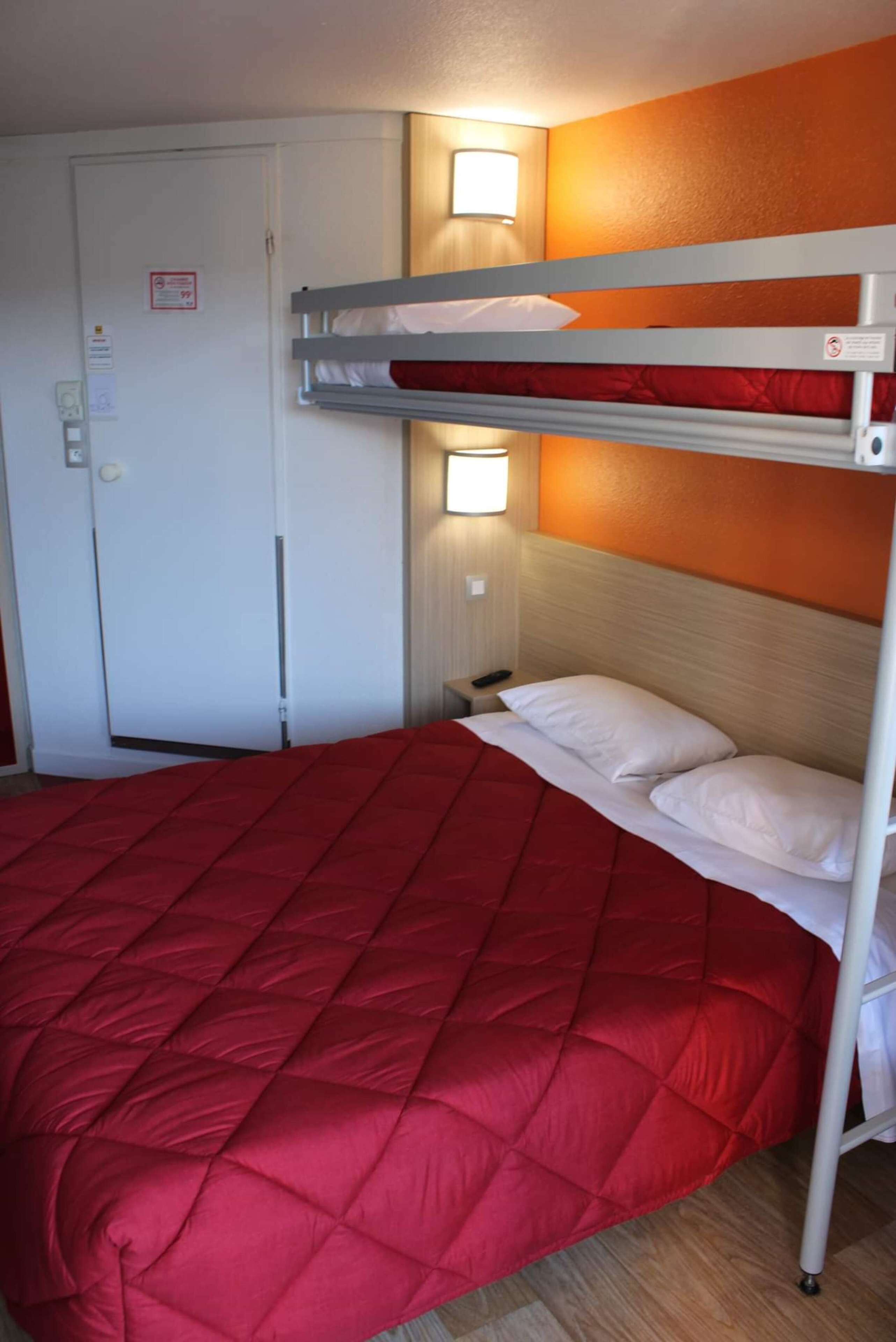 A bunk bed with a red quilted bedspread and white pillows, a nightstand with a lamp, and a door with a sign.