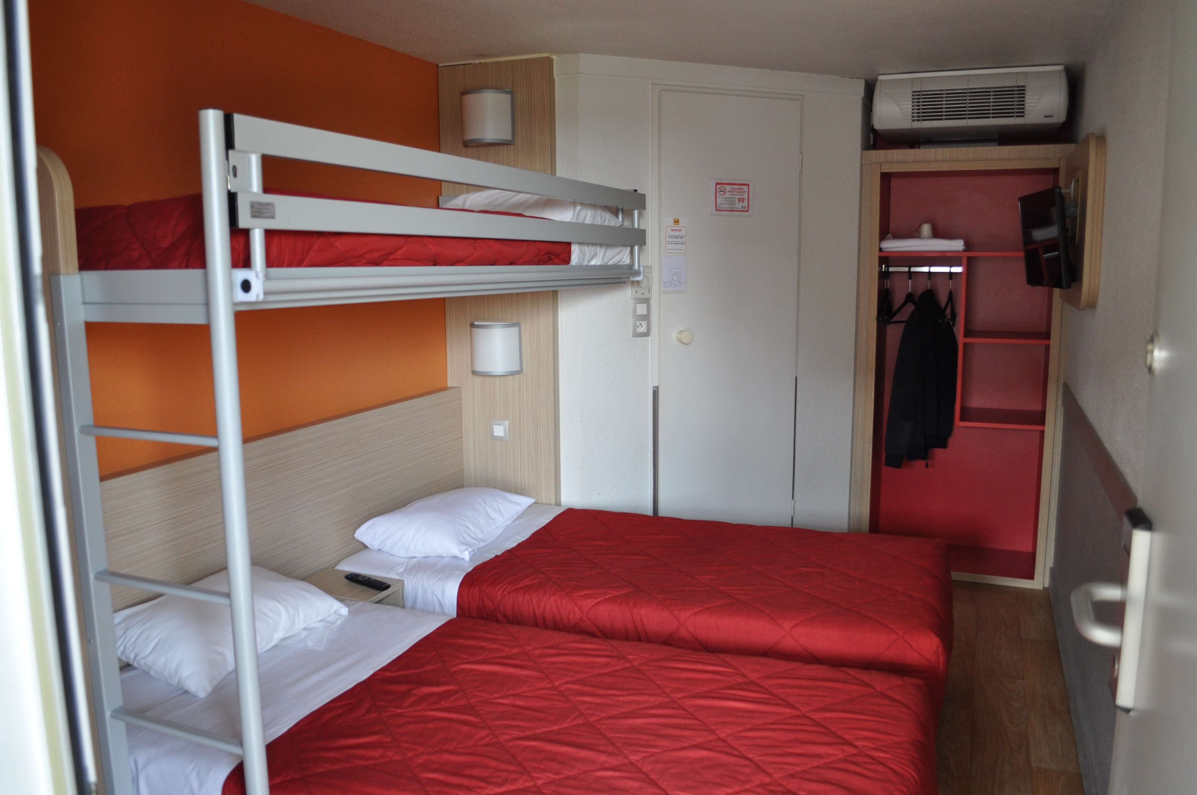 A bunk bed room with a red bedspread, a white wall, and a door with a coat rack.