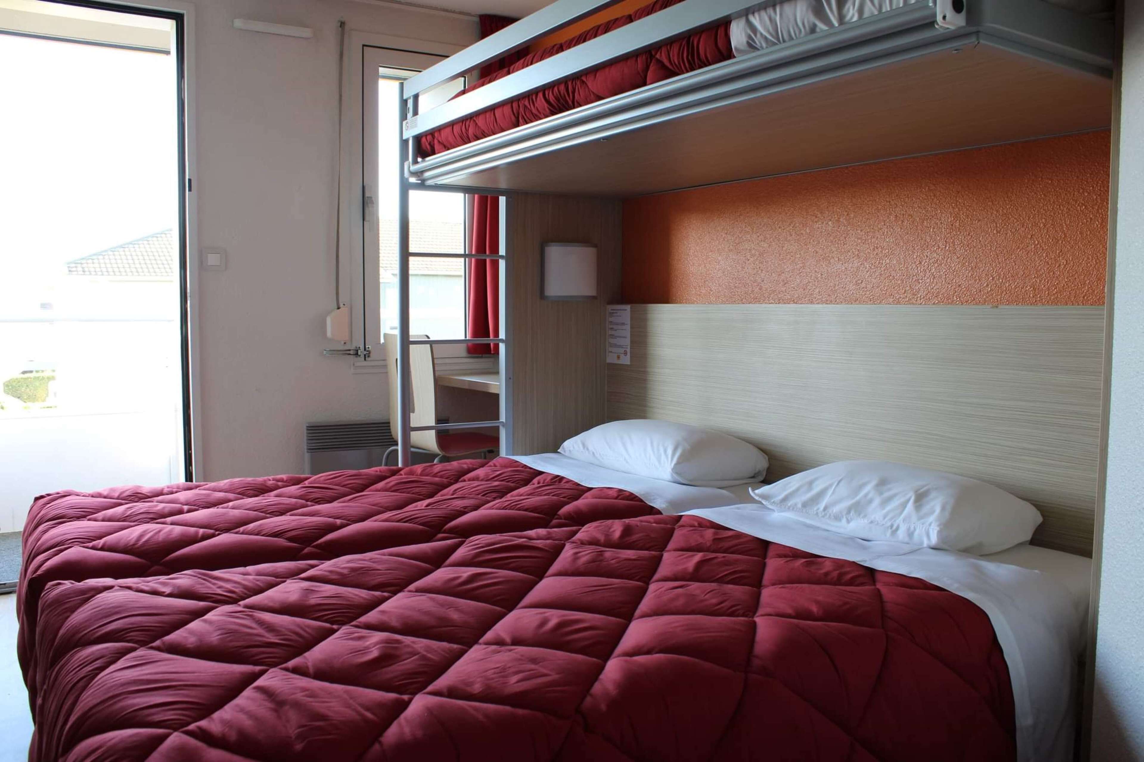 A single bed with a red quilt, a bunk bed above, a white headboard, and a window with red curtains.
