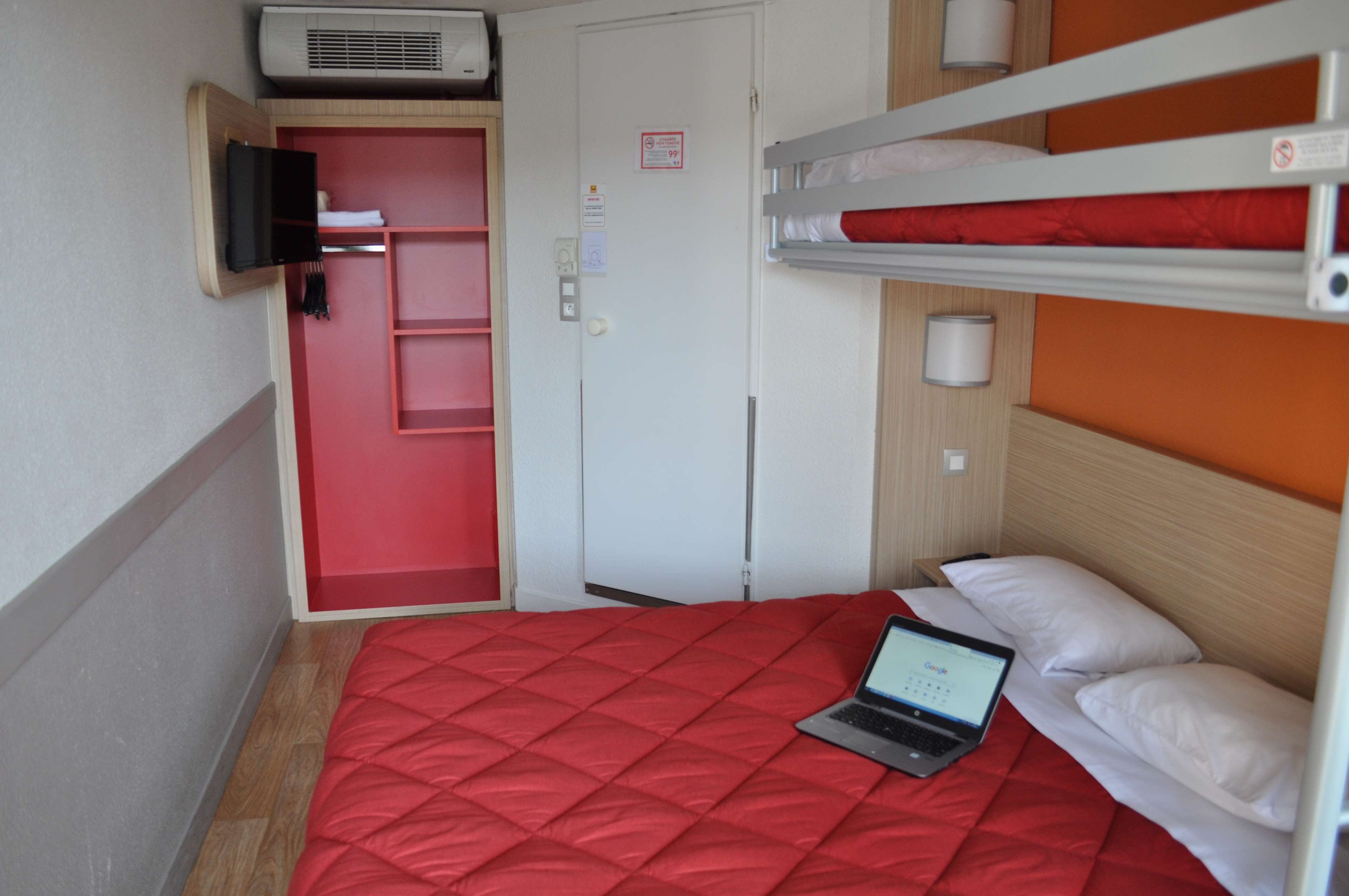 A bunk bed with a laptop on the lower bunk, a red bedspread, and a red door with a television mounted above it.