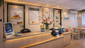 Un espace d’accueil d’hôtel avec un comptoir en bois, des affiches encadrées, un vase de fleurs et un coin salon avec des chaises et une table.