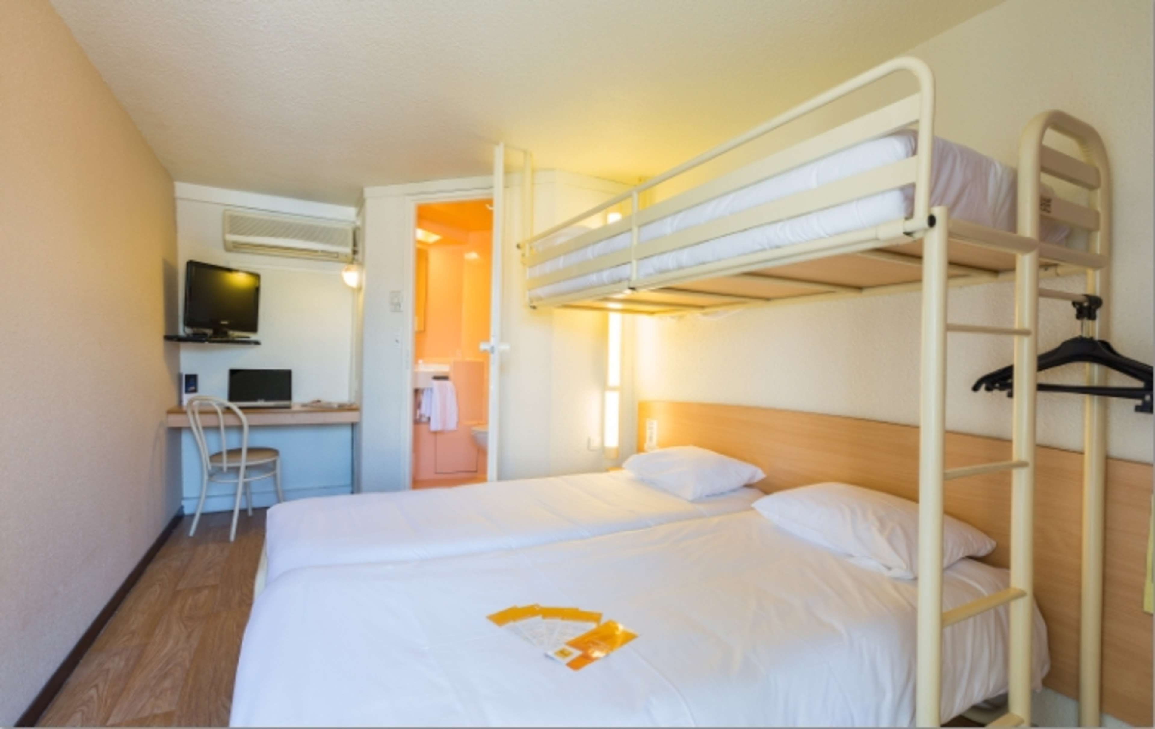 Une chambre avec un lit superposé, un bureau et une chaise, une télévision, et une salle de bain visible à travers une porte ouverte.