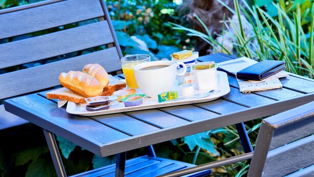 A breakfast tray with bread, butter, jam, a cup of coffee, and a book on a wooden table outdoors.