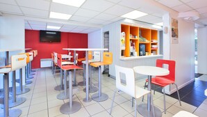A brightly lit cafeteria with multiple tables and chairs, a red wall with a television, and shelves with food items.