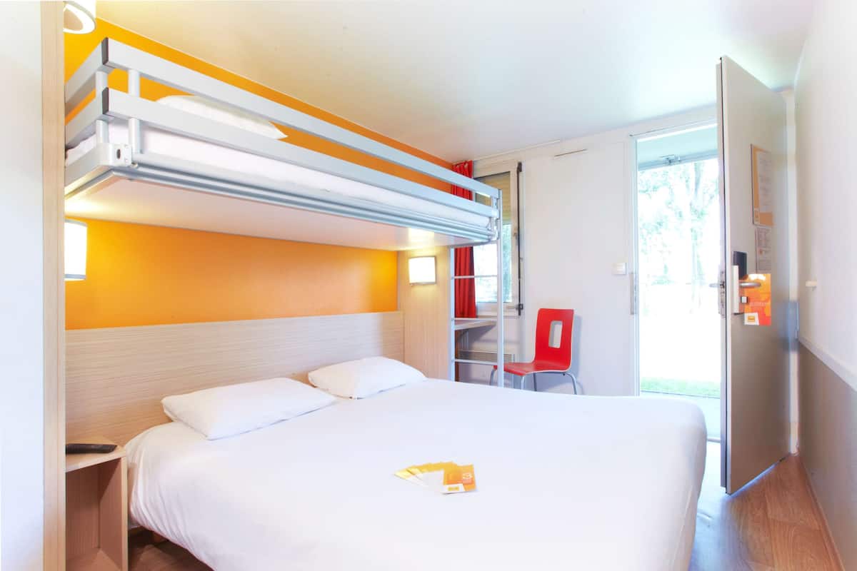 Een hotelkamer met een stapelbed, een eenpersoonsbed, een bureau met stoel en een deur die leidt naar een balkon.