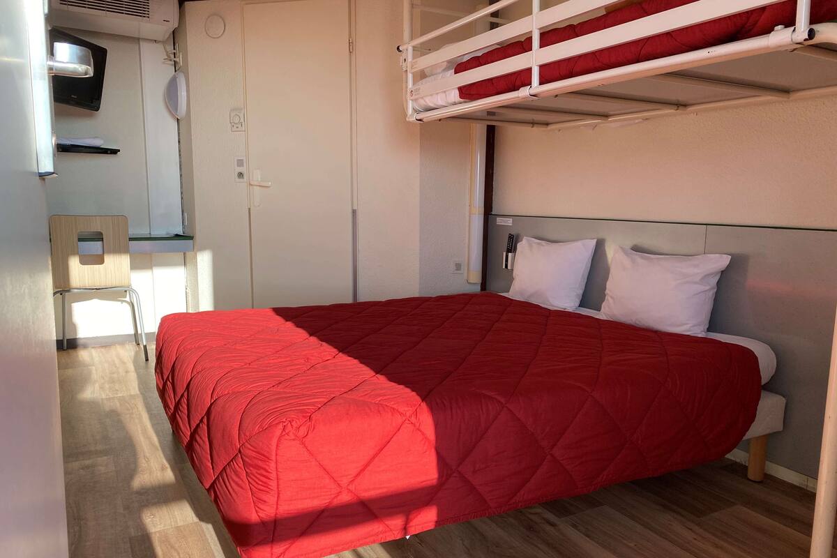 Habitación con una cama individual, colcha de patchwork roja, una cama litera arriba, una silla de madera y pared blanca con una puerta y un estante pequeño.