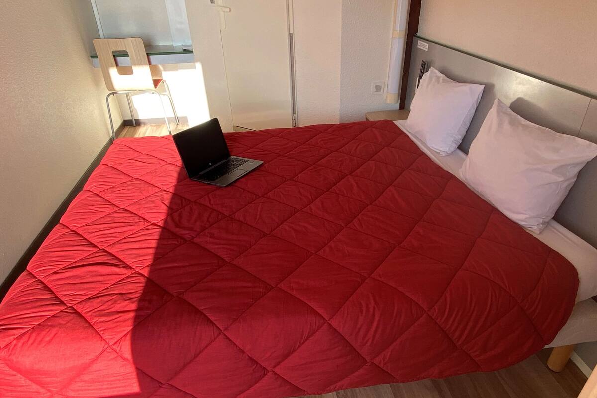 Una cama con un edredón rojo, una computadora portátil encima y almohadas blancas.
