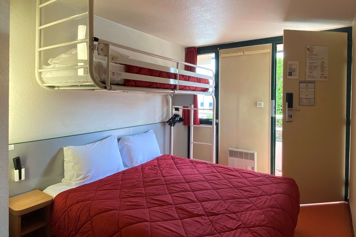 Habitación de hotel con literas, una cama individual con colcha roja, una mesita de noche de madera y una puerta que da a un balcón.