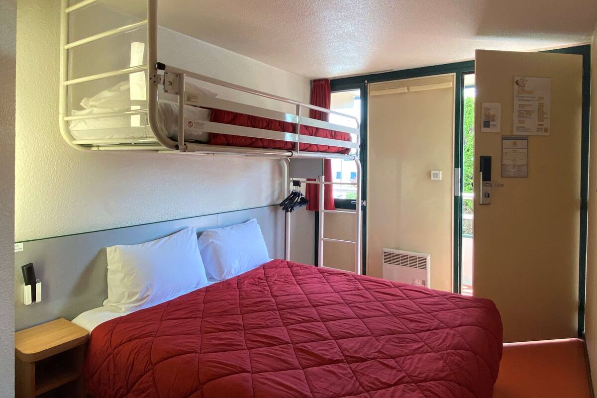 Habitación de hotel con literas, una cama individual con colcha roja, una mesita de noche de madera y una puerta que da a un espacio exterior.