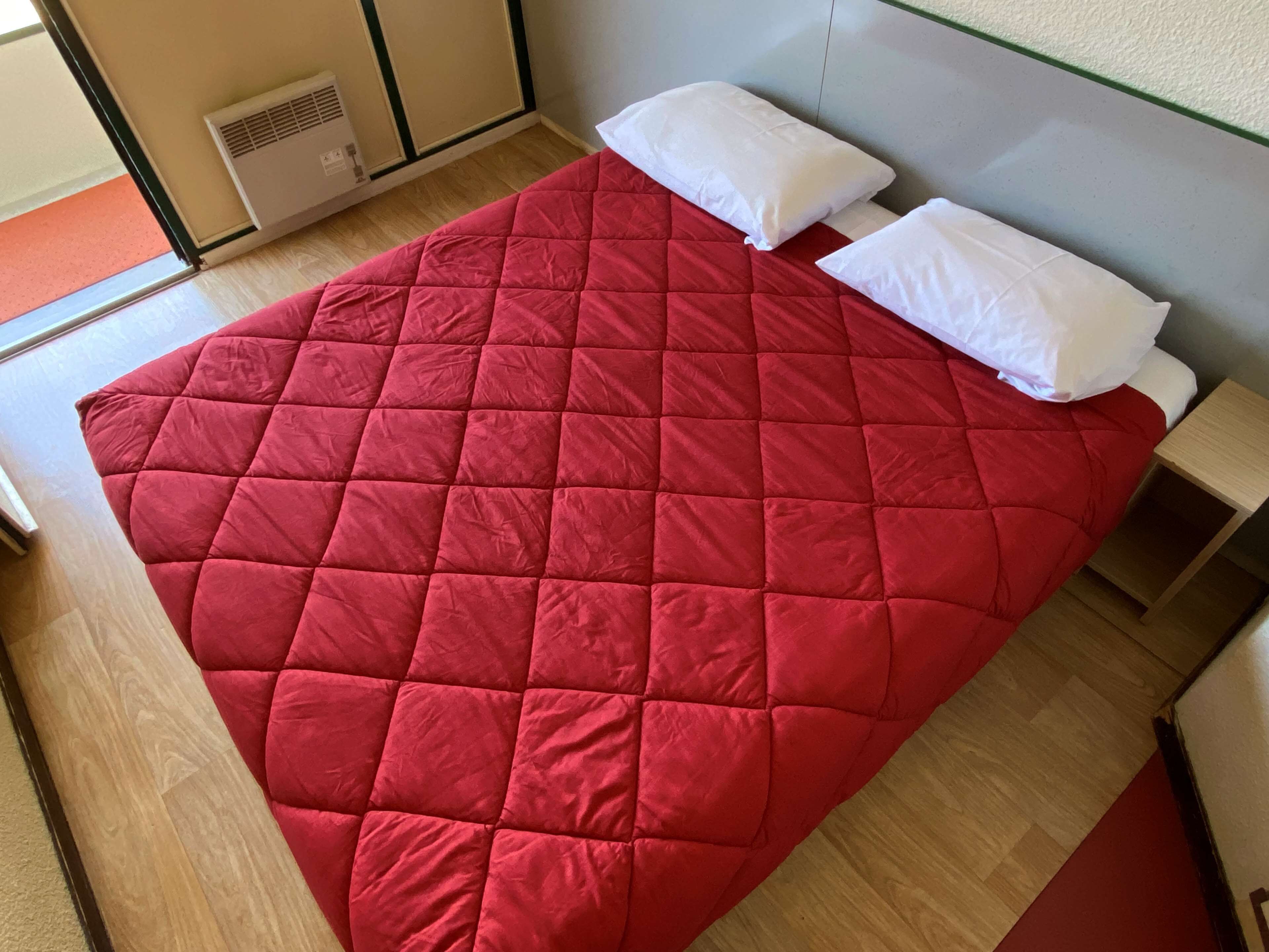 Een bed met een rode quilt en witte kussens in een kamer met houten vloer en een wit hoofdeinde.