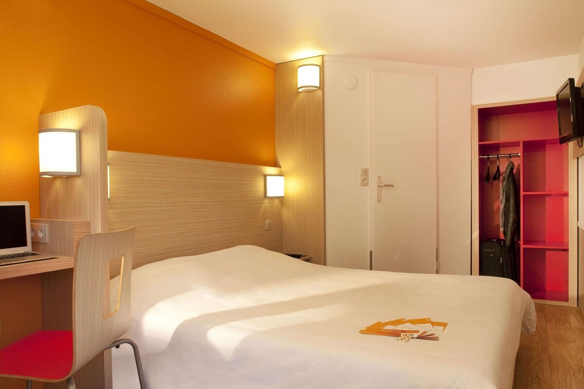A hotel room with a bed, a desk with a computer, a chair, a wardrobe, and a TV.