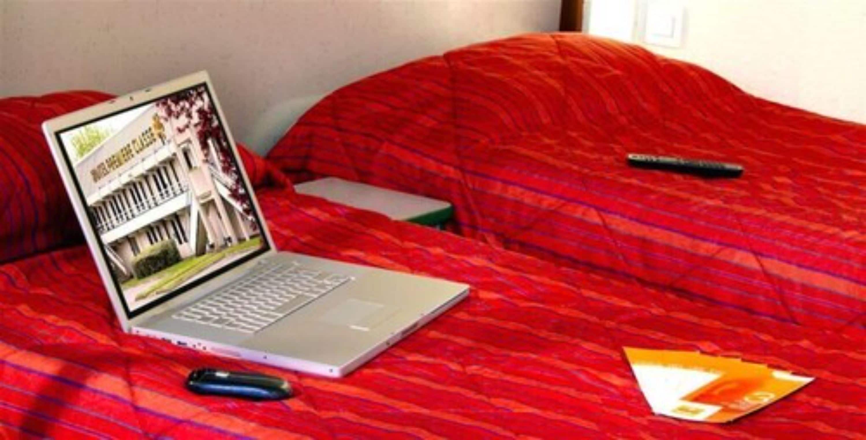 A laptop on a bed with a red blanket, a remote control, and a book.