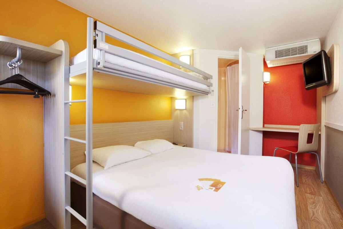 A hotel room with a bunk bed, a desk with a chair, a TV, and a wall-mounted air conditioner.