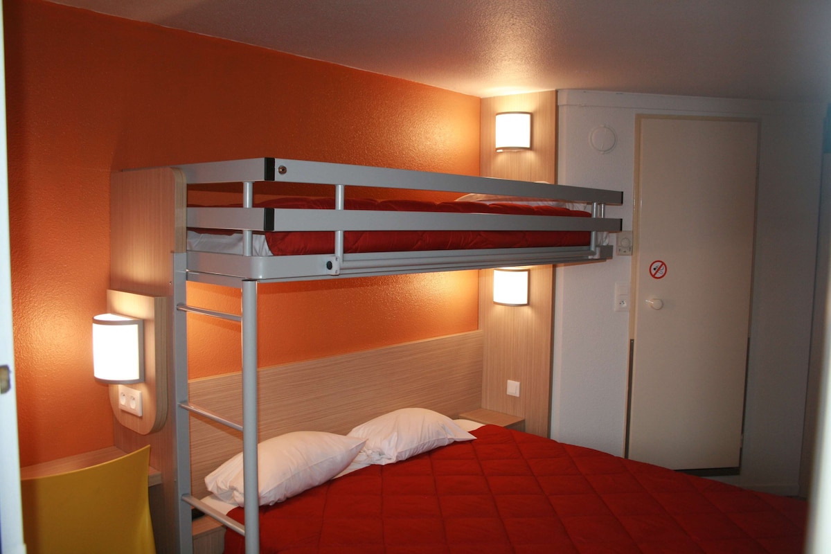 Una cama litera con colchón rojo, una mesita de noche con lámpara y una puerta con letrero de no fumar.