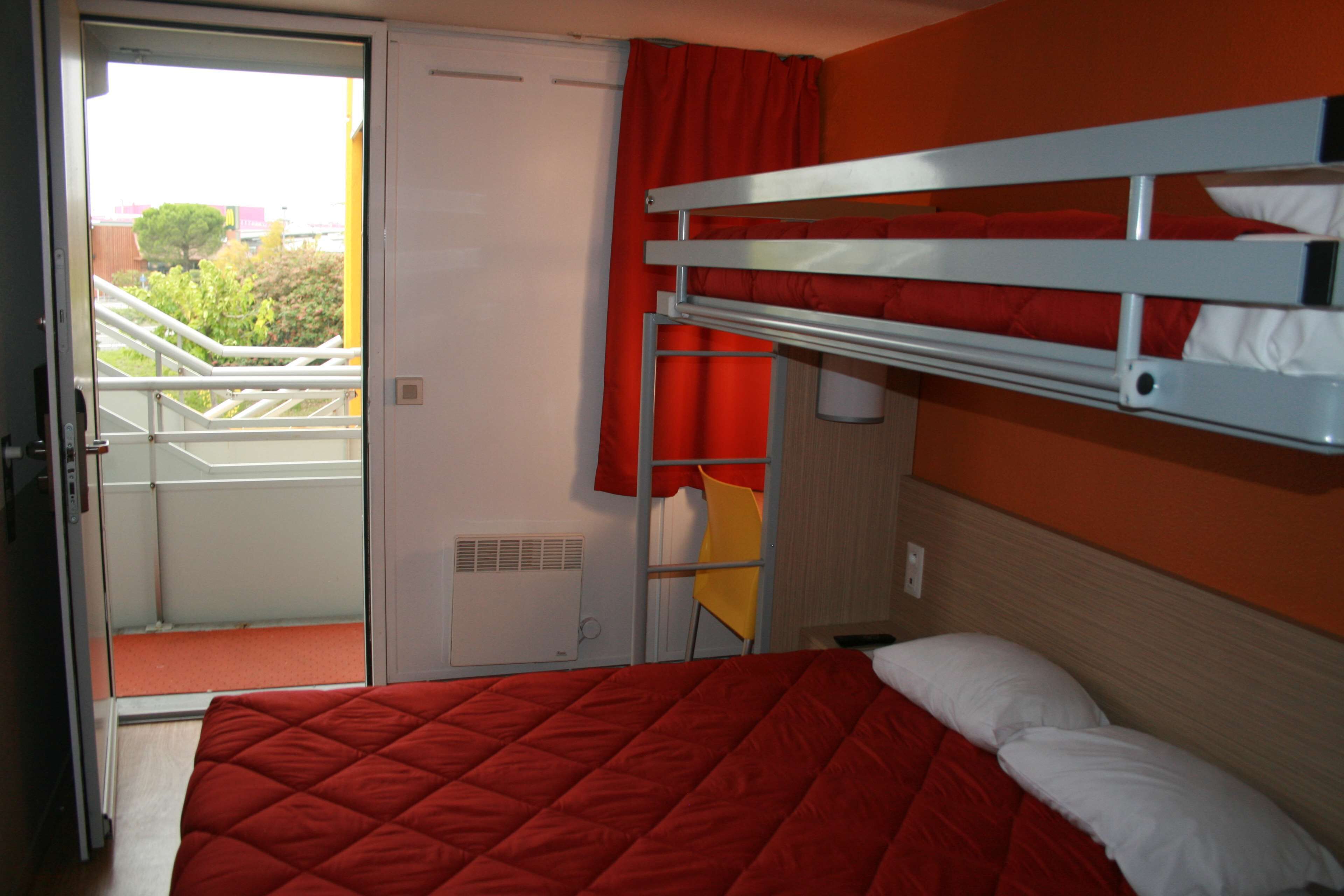 A room with a bunk bed, a red bedspread, a white pillow, a red curtain, and a yellow chair.