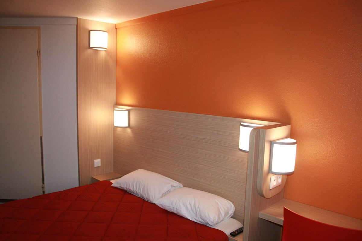 Habitación de hotel con colcha roja, almohada blanca y dos luces fijadas a la pared.