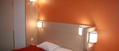 Habitación de hotel con colcha roja, almohada blanca y dos luces fijadas a la pared.