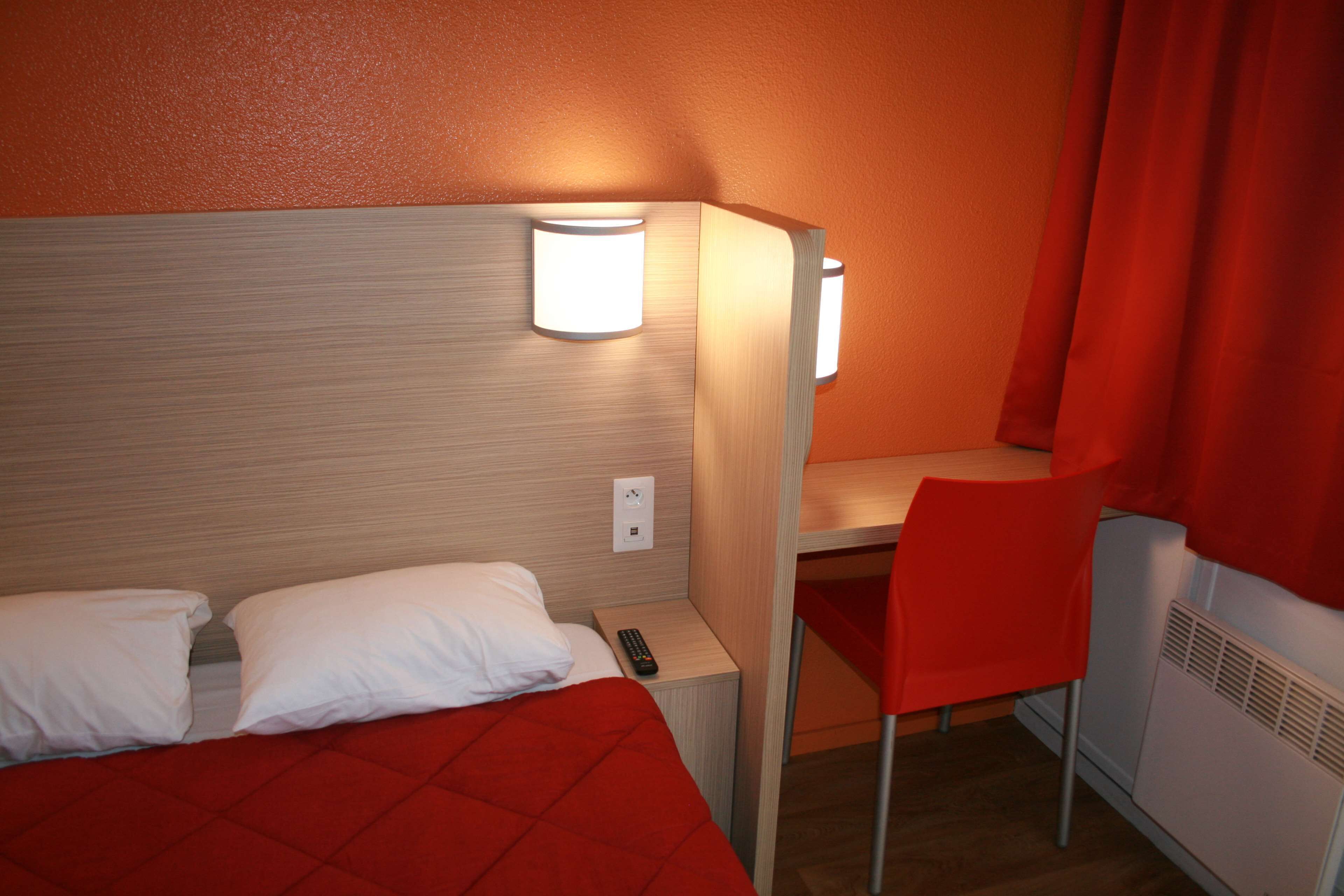 Une chambre d’hôtel avec un lit, un bureau et une chaise. Il y a une télécommande sur le bureau.