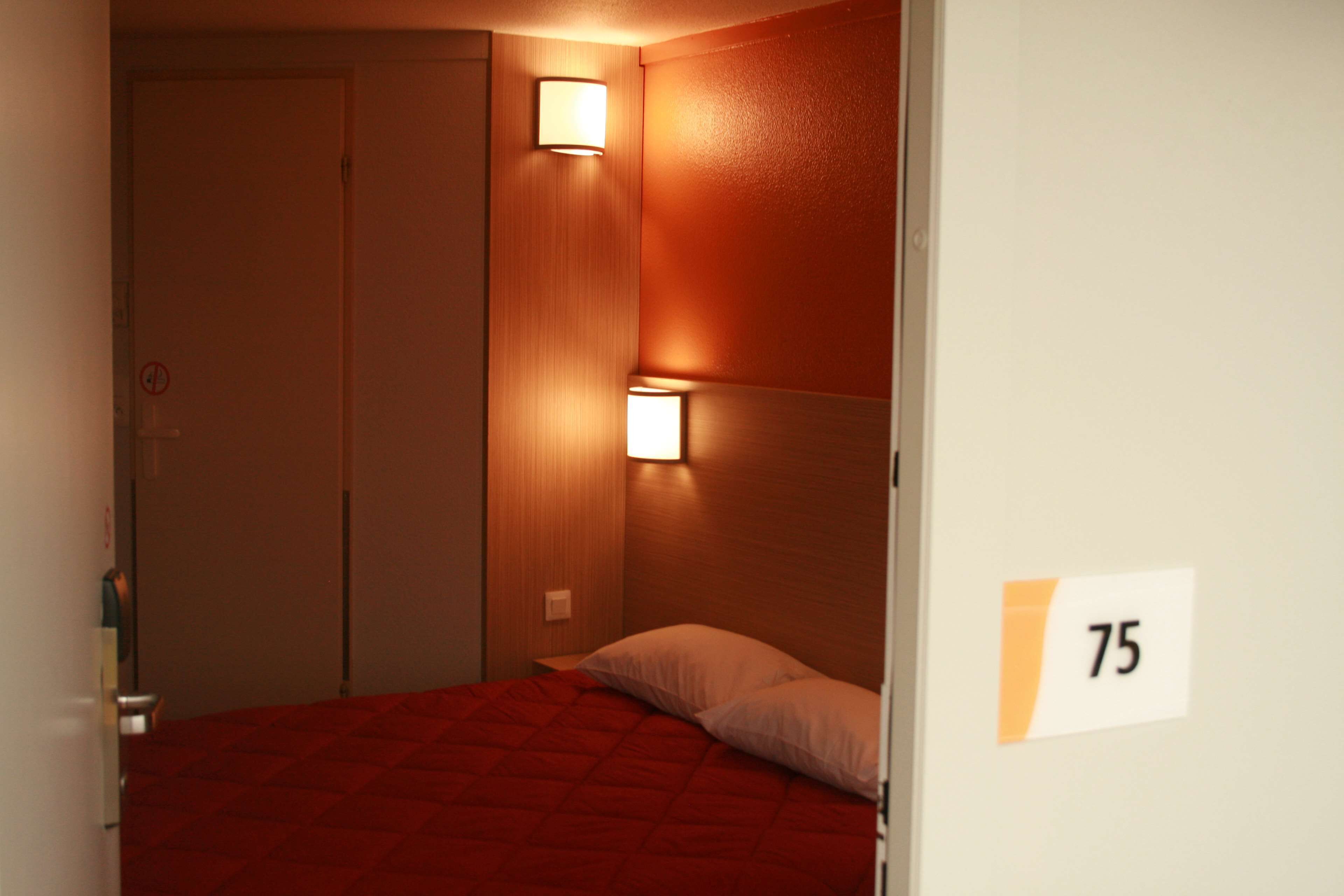 Habitación de hotel con una colcha roja, dos luces fijadas a la pared y una puerta con el número 75.