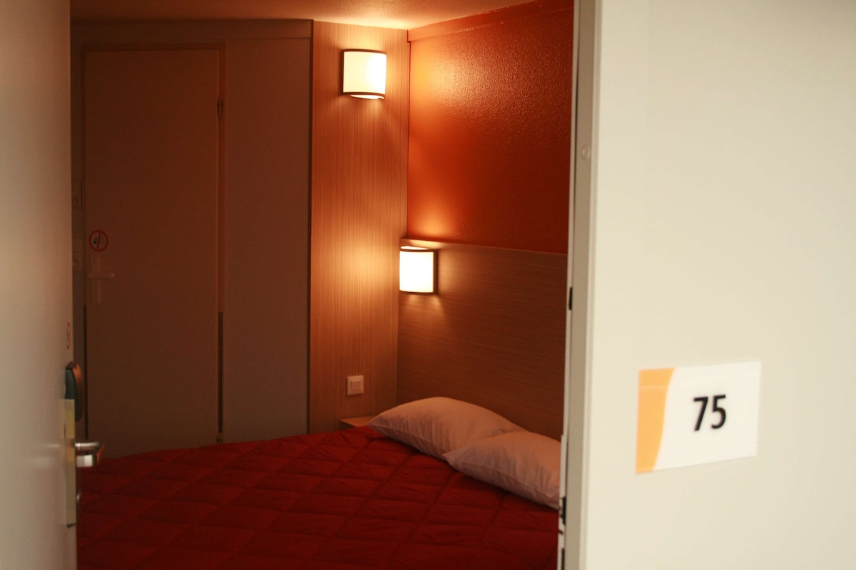 Habitación de hotel con una colcha roja, dos luces fijadas a la pared y una puerta con el número 75.