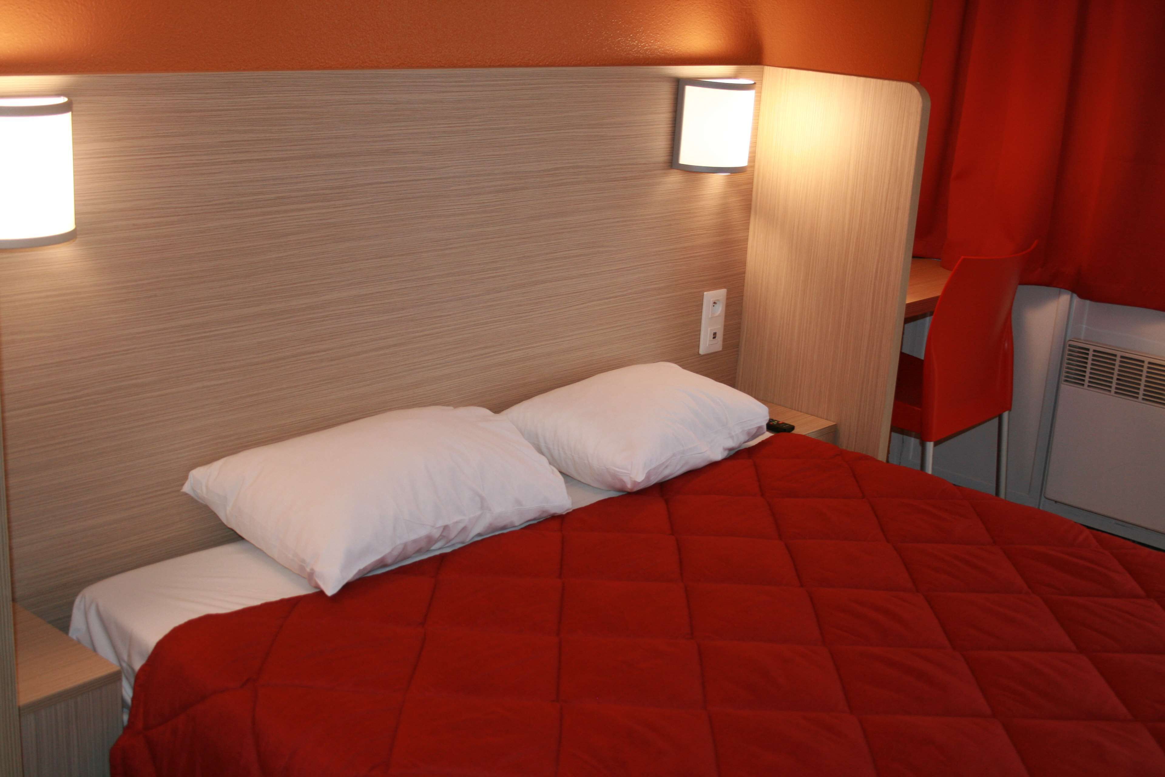 Una habitación de hotel con una cama, dos almohadas, una colcha roja y dos luces fijadas a la pared.
