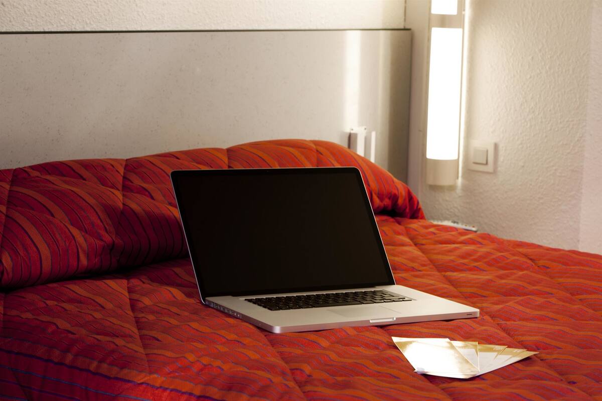Un ordinateur portable posé sur un lit, avec une couverture à motifs rouges et un oreiller blanc.