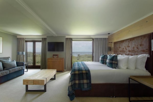 Habitación de hotel con cama, sofá, mesa de centro, televisión y balcón con vistas.