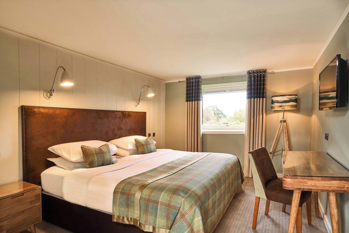 A hotel room with a large bed, a desk, a chair, a TV, and a window with curtains.