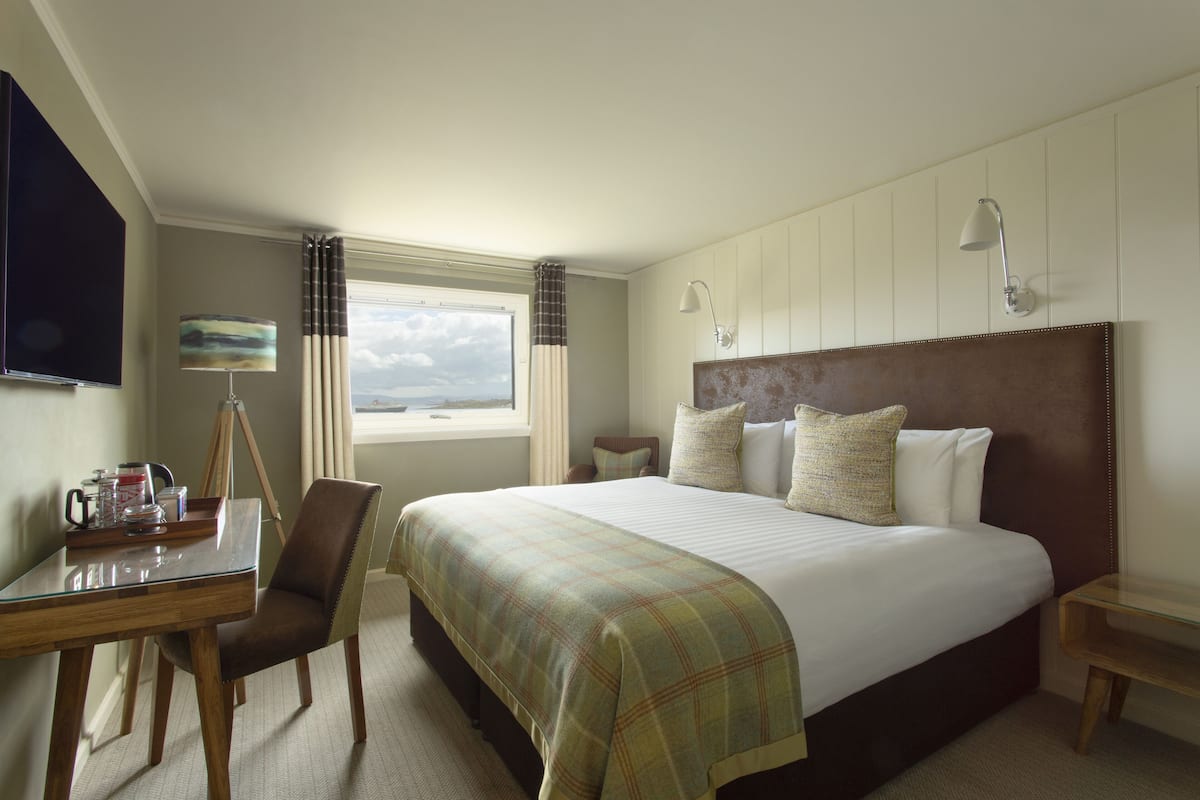 A hotel room with a large bed, a desk with a chair, a lamp, and a window with curtains.