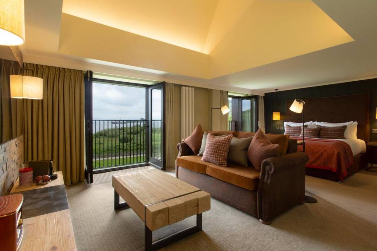 A hotel room with a sofa, a wooden coffee table, a bed with a headboard, and a view of the outdoors through a large window.