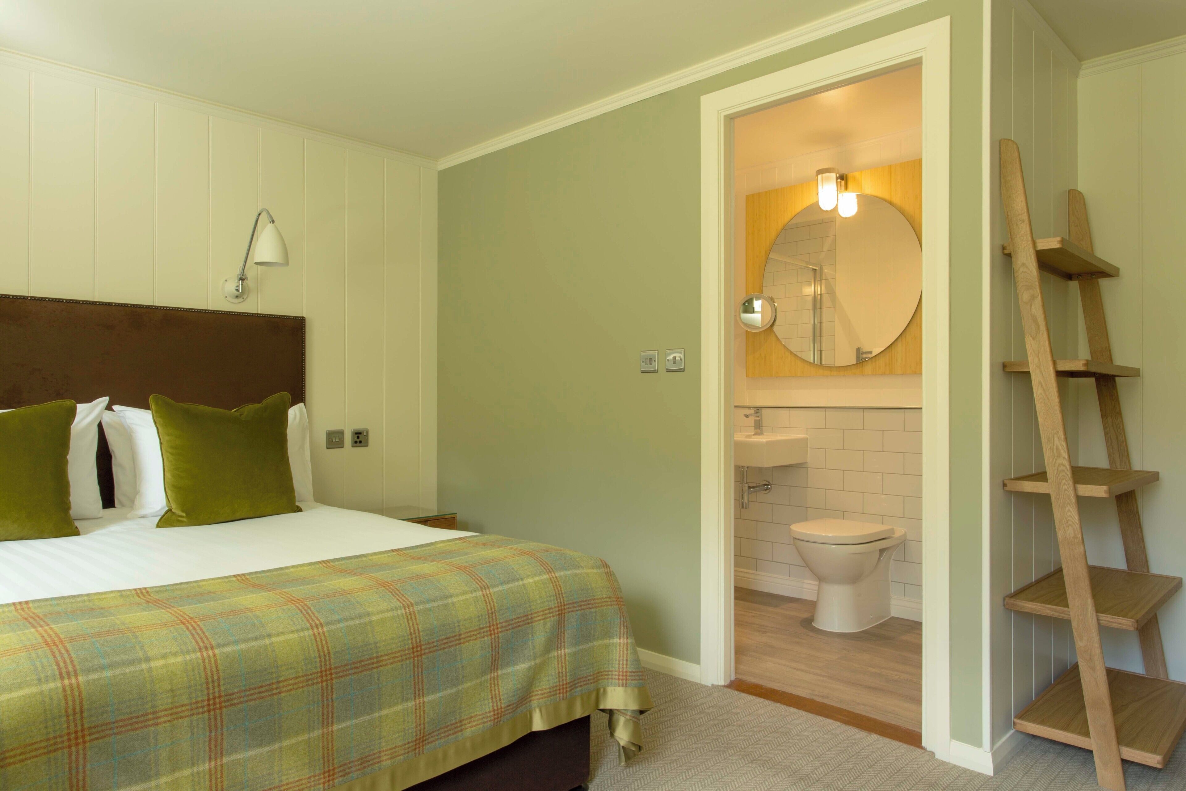 Une chambre à coucher avec un lit, des oreillers, une lampe de chevet, et une salle de bain équipée de toilettes, d’un lavabo, d’un miroir et d’une échelle en bois.