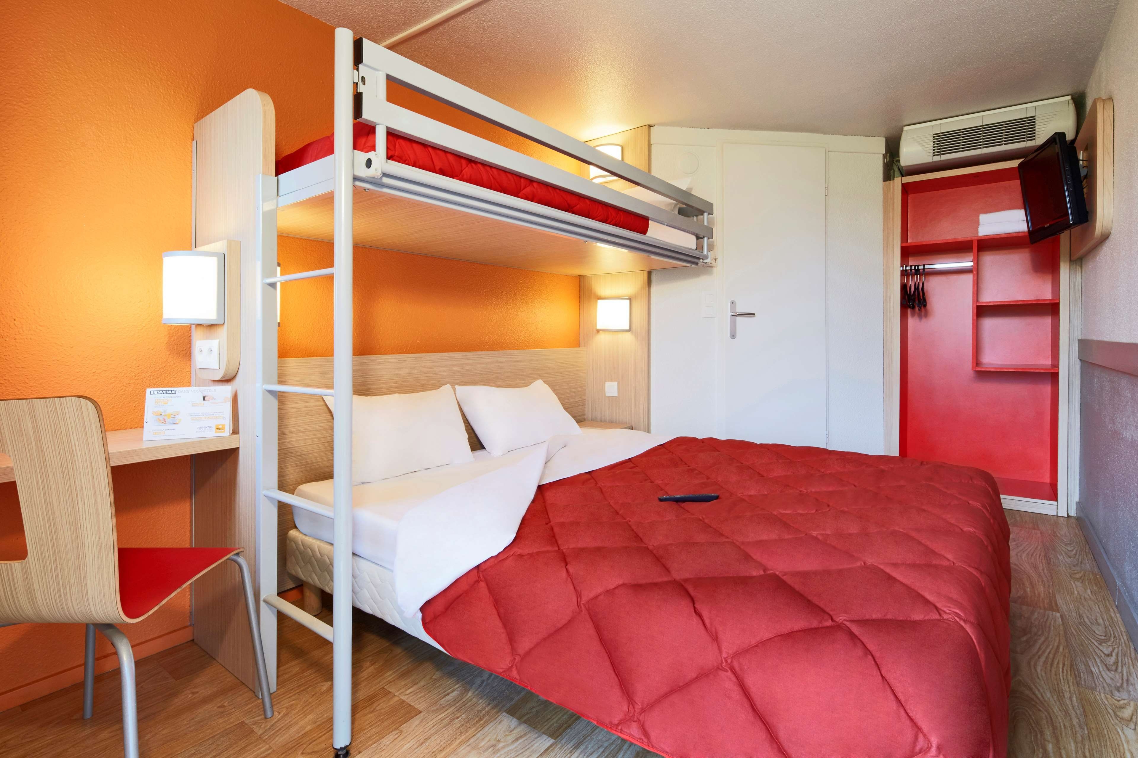 A bunk bed with a red quilt, a desk with a lamp, and a wardrobe with a red door.