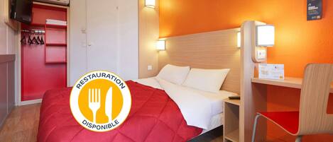 A hotel room with a bed, a desk, a chair, and a wardrobe. There is a sign indicating a restaurant is available.