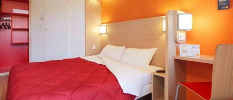 Habitación de hotel con cama, escritorio, silla, armario y una lámpara fijada a la pared.