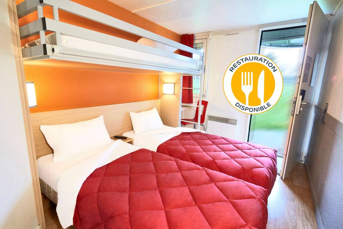 Une chambre d’hôtel avec un lit superposé, une couvre-lit rouge matelassée, une table de chevet, une lampe et une porte coulissante donnant sur l’extérieur.