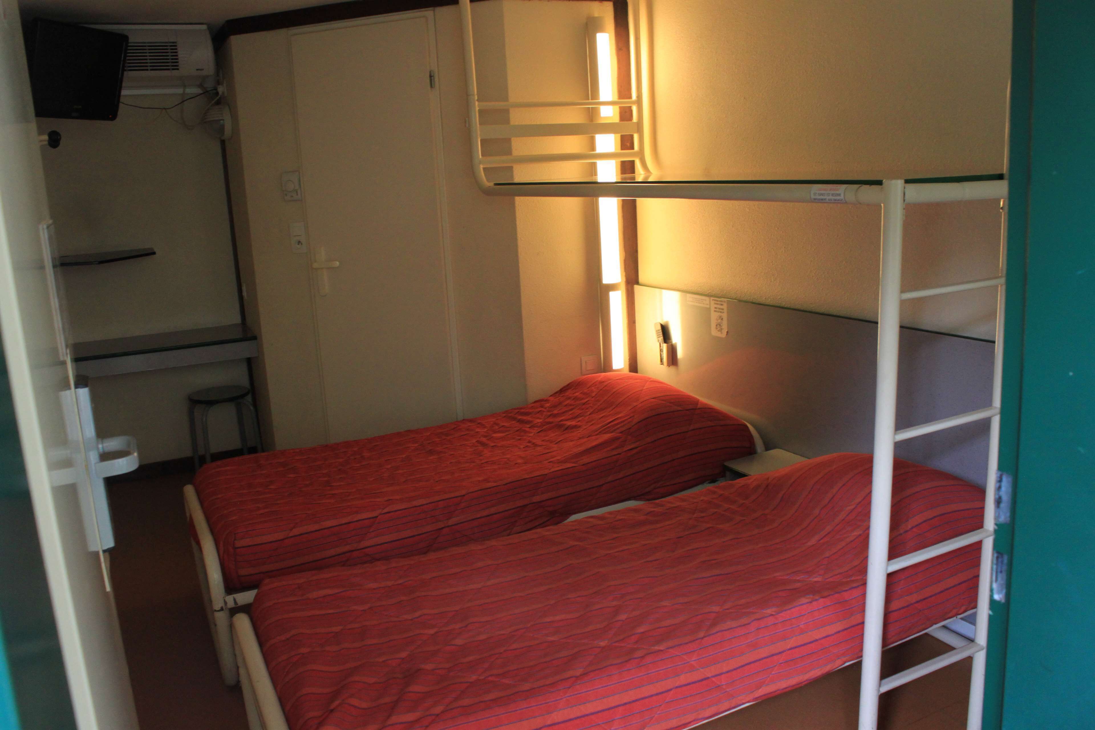 A bunk bed room with a single bed and a bunk above. There is a small table and a chair, a wall-mounted heater, and a television on the wall.