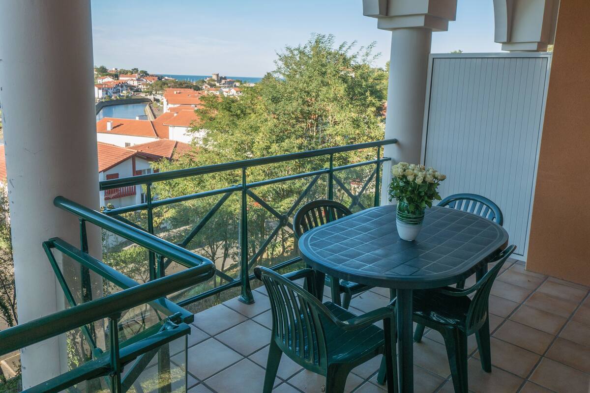 Ein Balkon mit rundem Tisch und Stühlen, einem Blumentopf und Blick auf eine Stadt mit roten Dächern.