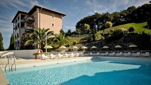 A swimming pool with lounge chairs and umbrellas, surrounded by a stone wall and a building with balconies. - Résidence Soko-Eder (Ciboure)