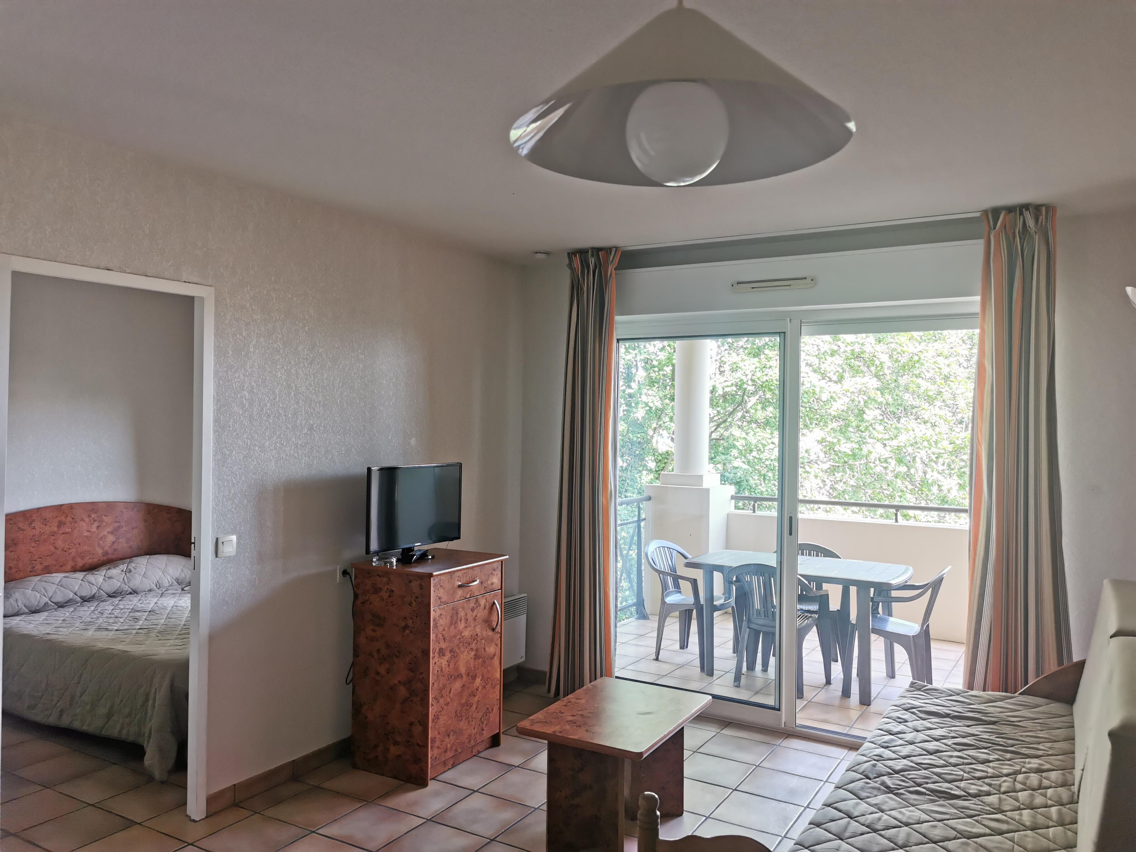 Une chambre d’hôtel avec un lit, un téléviseur posé sur une commode en bois, une petite table et un balcon équipé de chaises et d’une table.