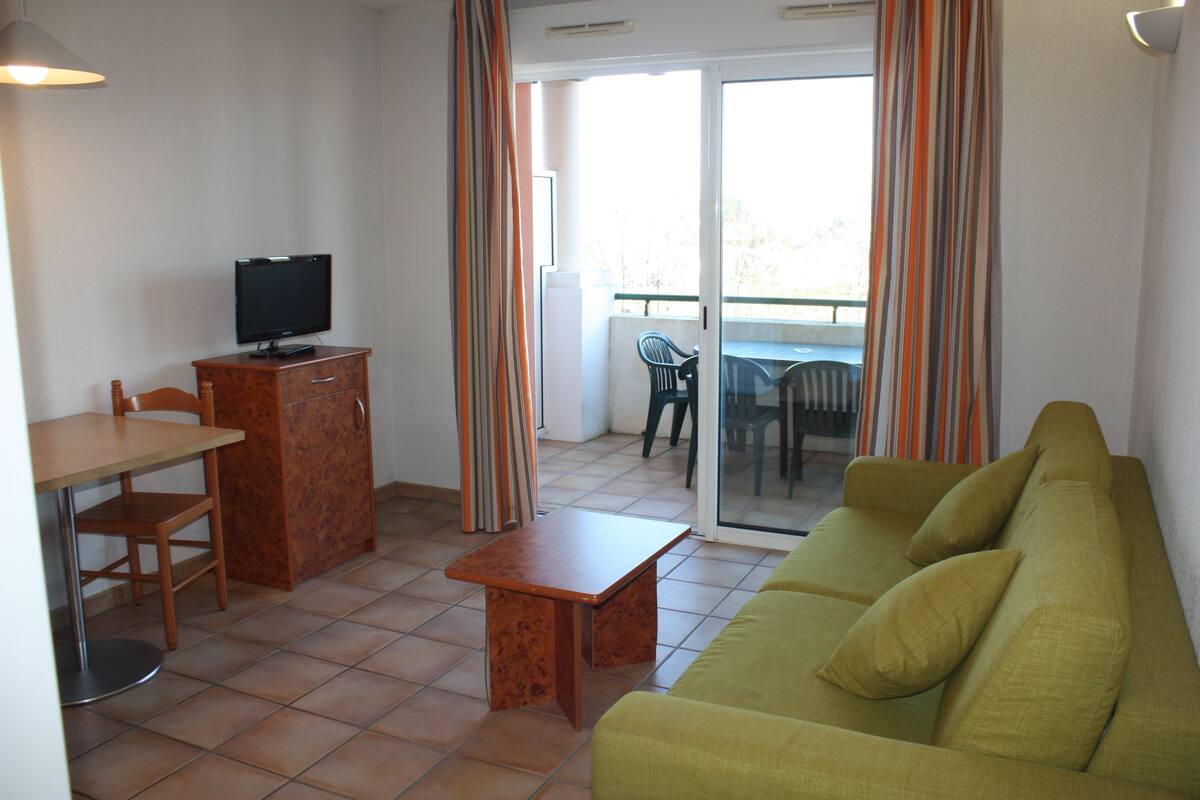 Ein kompakter Wohnraum mit Sofa, Fernseher auf einem Schrank, Esstisch und Balkon mit einem Stuhl.