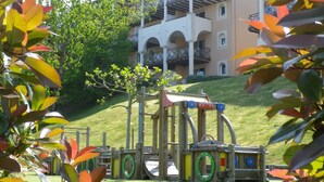 A playground with a wooden structure, surrounded by green grass and trees. In the background, there is a multi-story building with balconies. - Résidence Soko-Eder (Ciboure)
